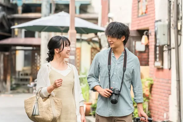 若いカップルが笑顔で街並みを歩いています。男性はカメラを首から下げ、スーツケースを引いており、旅行中のようです。