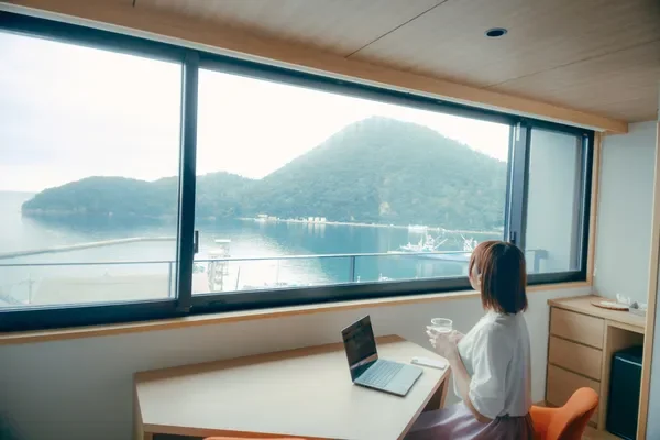 窓から広がる美しい海の景色を眺めながら、女性がノートパソコンに向かっています。グラスを片手にリラックスした様子で、仕事や休憩をしているようです。開放的な空間で穏やかな時間が流れています。