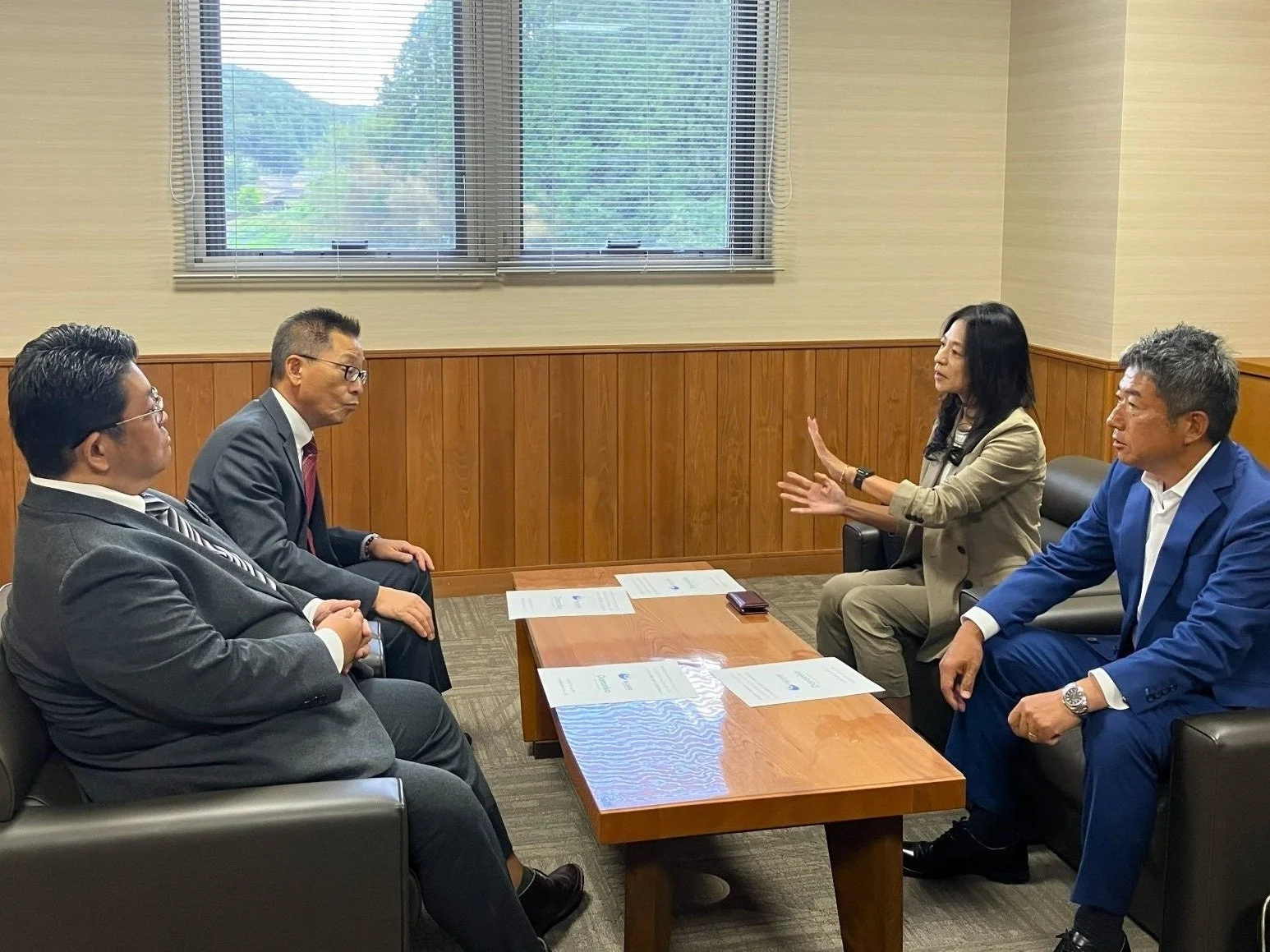 会議室のような場所で、4人の男女が木製のテーブルを囲んで座り、活発に話し合っている様子。