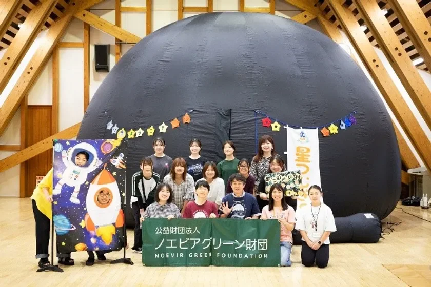移動式プラネタリウムの前で、宇宙テーマの顔出しパネルを楽しむ若者たちが集合写真を撮っている。公益財団法人ノエビアグリーン財団による教育イベントの様子。