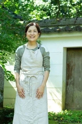 屋外の緑豊かな場所で、エプロンを着用した女性が穏やかな笑顔でカメラを見ている。自然な光の中で、和やかな雰囲気が感じられる一枚。