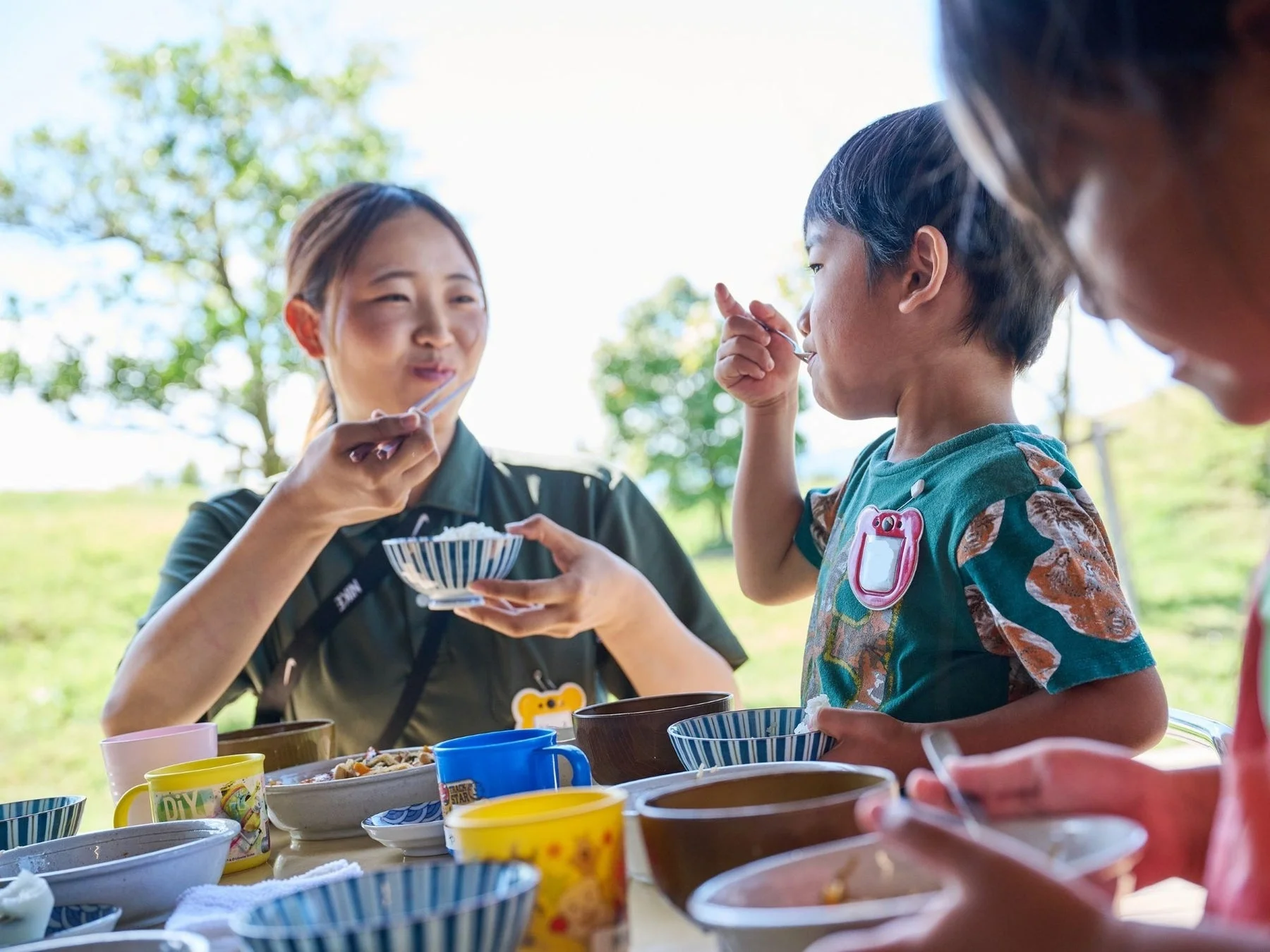 晴れた日の屋外で、大人と子供たちが一緒に食事を楽しんでいます。