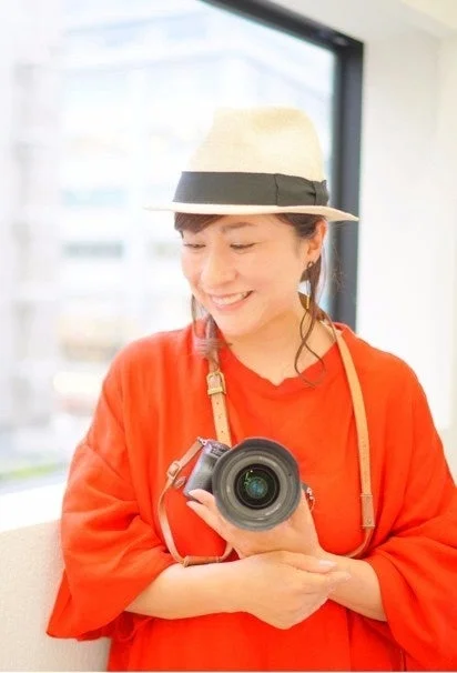 白いハットとオレンジ色のトップスを着用した女性が、笑顔でカメラを手にしているポートレート写真です。窓際のような明るい場所で撮影されており、リラックスした雰囲気が伝わります。